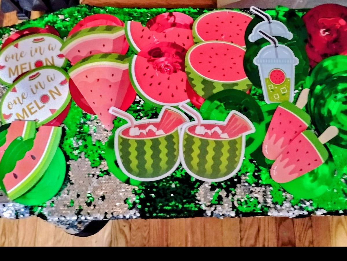 A collection of watermelon-themed party decorations, including paper cutouts of watermelon slices, popsicles, limes, and drinks, along with round signs that read 'one in a MELON one in a MELON', arranged on a black sequined backdrop over a wooden floor.